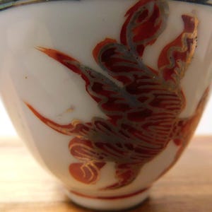 May include: White ceramic cup with a flared shape, adorned with a red and gold bird design. The bird's wings are spread, and the cup has a blue and black rim. The cup is likely part of a tea set or decorative item.