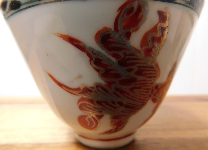 May include: White ceramic cup with a flared shape, adorned with a red and gold bird design. The bird's wings are spread, and the cup has a blue and black rim. The cup is likely part of a tea set or decorative item.