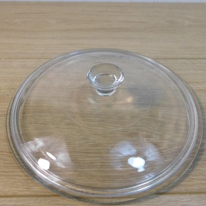 May include: A clear glass lid with a small, rounded handle. The lid is circular and appears to be for a pot or pan. The glass is transparent, allowing a view of the contents below. The lid is sitting on a light brown wooden surface.