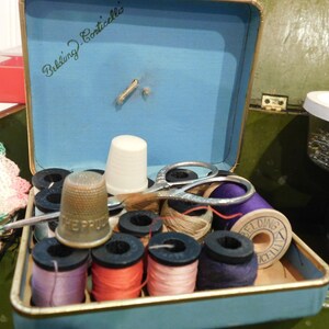 May include: Open blue sewing kit with various sewing supplies. Includes spools of thread in multiple colors, a thimble, scissors, and a white thimble. The kit is labeled "Belding-Corticelli" in cursive.