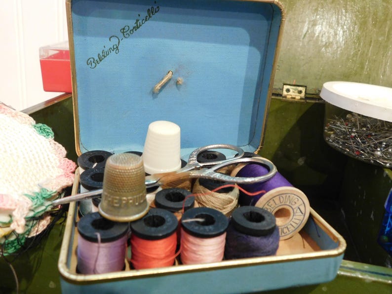 May include: Open blue sewing kit with various sewing supplies. Includes spools of thread in multiple colors, a thimble, scissors, and a white thimble. The kit is labeled "Belding-Corticelli" in cursive.