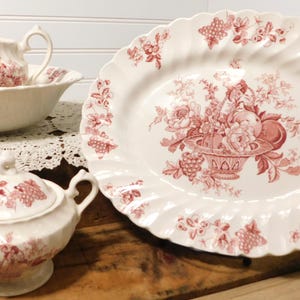 May include: A cream-colored ceramic set featuring a large oval platter, a lidded sugar bowl with two handles, and a creamer pitcher. Each piece is decorated with a red floral and fruit transferware pattern, including scalloped edges on the platter.