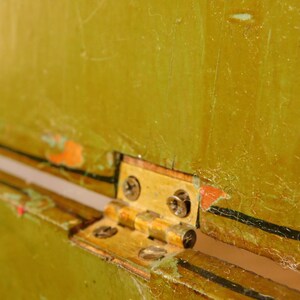 May include: Close-up of a brass hinge attached to a distressed, olive-green painted surface. The hinge is secured with small screws. The paint shows signs of wear and tear, with visible scratches and areas where the paint has chipped away.