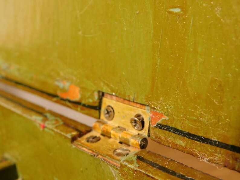 May include: Close-up of a brass hinge attached to a distressed, olive-green painted surface. The hinge is secured with small screws. The paint shows signs of wear and tear, with visible scratches and areas where the paint has chipped away.
