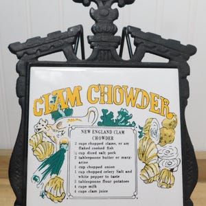 May include: A decorative black metal trivet with a white tile featuring a recipe for New England Clam Chowder. The tile has yellow and green illustrations of clams and vegetables, with the words "CLAM CHOWDER" in large yellow letters.