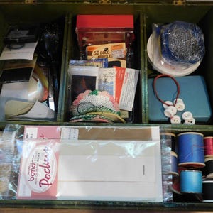 May include: An open, vintage wooden sewing box with multiple compartments. The box contains spools of thread, needles, buttons, pins, and other sewing notions. The box is painted a dark green color.