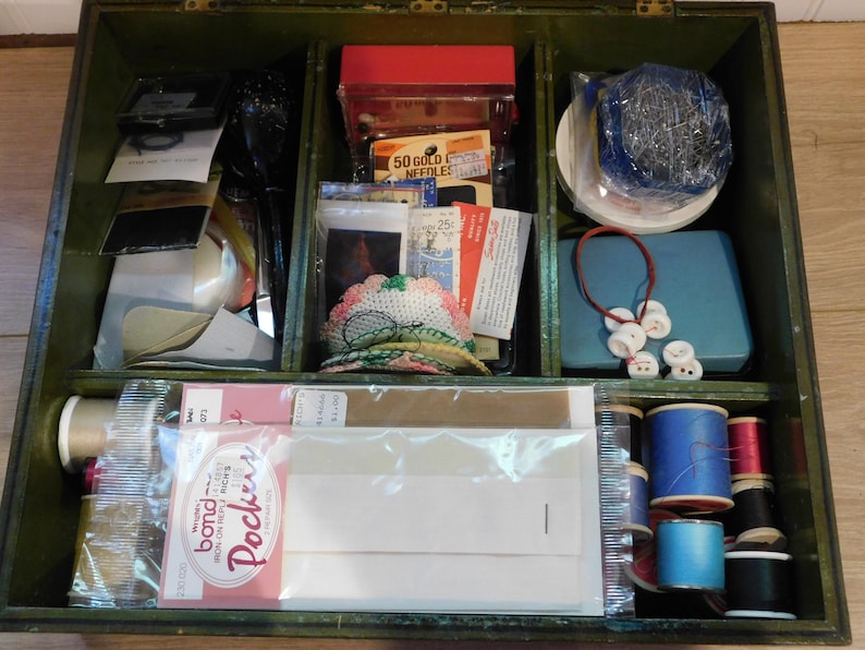 May include: An open, vintage wooden sewing box with multiple compartments. The box contains spools of thread, needles, buttons, pins, and other sewing notions. The box is painted a dark green color.