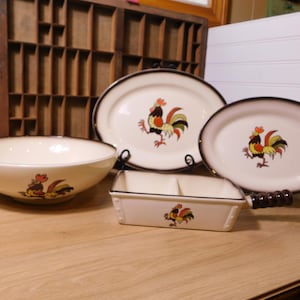 May include: A collection of vintage ceramic serving dishes with a rooster motif. The set includes a large bowl, two oval platters, and a divided rectangular dish. Each piece has a cream-colored base, a brown rim, and a colorful rooster illustration.