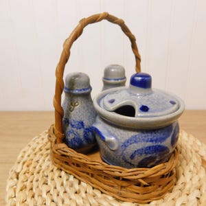 May include: A ceramic salt and pepper set and a lidded container with a spoon rest, all in a woven basket. The ceramic pieces are gray with blue swirl designs. The basket has a woven handle.