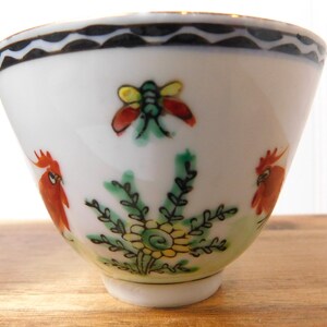 May include: White ceramic bowl with hand-painted floral and insect designs. The bowl features a black wavy border, a red and yellow insect, two red rooster figures, and a green and yellow floral design.