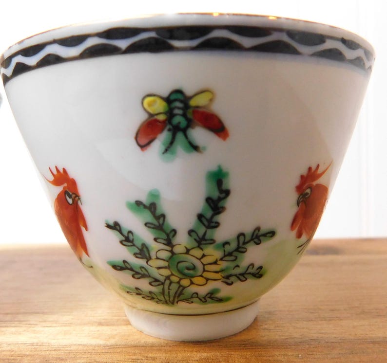May include: White ceramic bowl with hand-painted floral and insect designs. The bowl features a black wavy border, a red and yellow insect, two red rooster figures, and a green and yellow floral design.