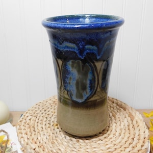 May include: A handcrafted ceramic vase with a flared top. The vase features a gradient of colors, including a deep blue rim, flowing into shades of blue and green, and transitioning to a tan base. The vase is placed on a woven mat.