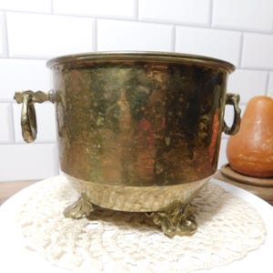 May include: A round brass planter with a weathered patina, featuring two side ring handles and three ornate, decorative feet. The metal surface shows variations in colour and texture, with a wide opening and a sturdy, bowl-shaped body.