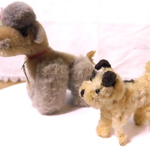 May include: Two vintage stuffed dog toys. One is larger, with tan and gray fur, a red collar, and a gray pom-pom head. The smaller dog is tan with black spots and a black patch on its head. Both have black eyes and noses.