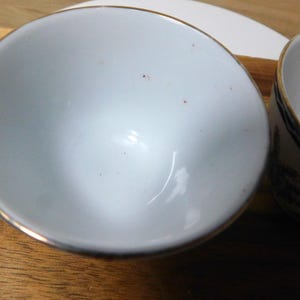 May include: Two white ceramic bowls with gold trim are set on a wooden surface. One bowl is in the foreground, and the other is partially visible in the background. The bowls appear to be empty.