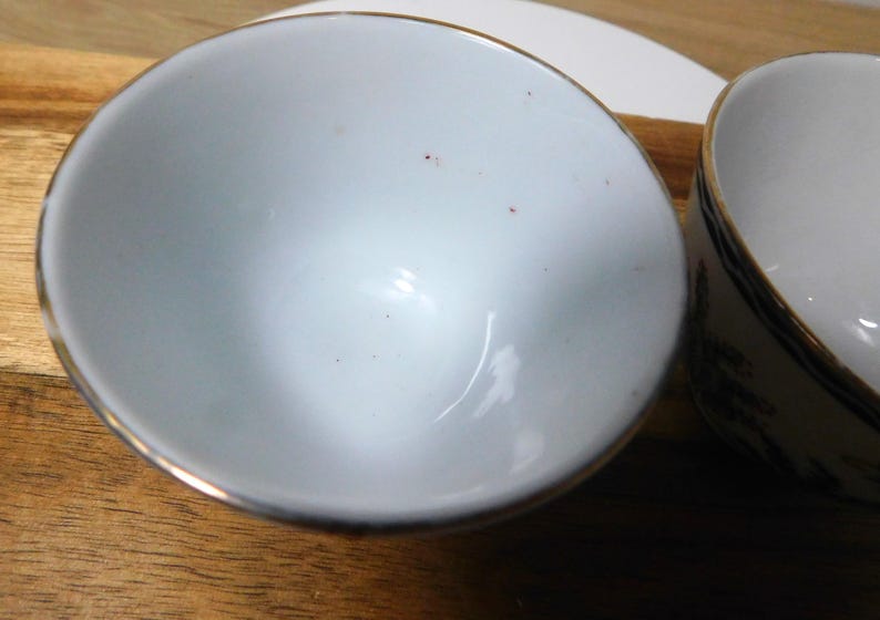 May include: Two white ceramic bowls with gold trim are set on a wooden surface. One bowl is in the foreground, and the other is partially visible in the background. The bowls appear to be empty.