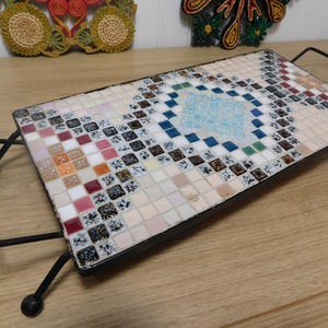 May include: Rectangular mosaic tile serving tray with a black metal frame and handles. The mosaic features a geometric pattern with squares in shades of white, blue, red, and brown. The tray is supported by four curved legs.