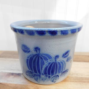May include: A blue and gray ceramic pot with a pumpkin design. The pot has a rounded shape with a blue rim and a pumpkin and leaf pattern in blue. The pot is sitting on a wooden surface.