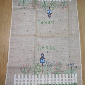 May include: A beige linen kitchen towel with a printed design featuring a white picket fence, flowers, and birdhouses. The word "HERBS" is printed in green. The towel has a green and white border.