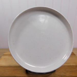May include: A round, off-white ceramic plate with a speckled surface. The plate has a slightly raised rim and is displayed on a wooden surface. The plate is approximately 12 inches in diameter. The plate is a neutral color and can be used for serving food.