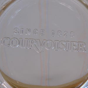 Pode incluir: Close-up de uma superf&iacute;cie de vidro transparente com o texto em relevo "SINCE 1828 COURVOISIER". O vidro tem um padr&atilde;o de diamante na borda e um ligeiro tom &acirc;mbar.