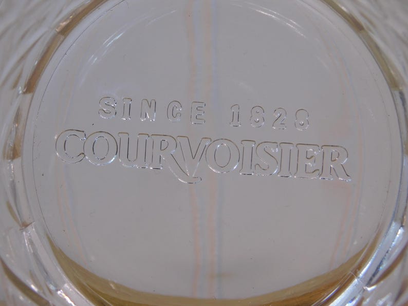 Pode incluir: Close-up de uma superf&iacute;cie de vidro transparente com o texto em relevo "SINCE 1828 COURVOISIER". O vidro tem um padr&atilde;o de diamante na borda e um ligeiro tom &acirc;mbar.