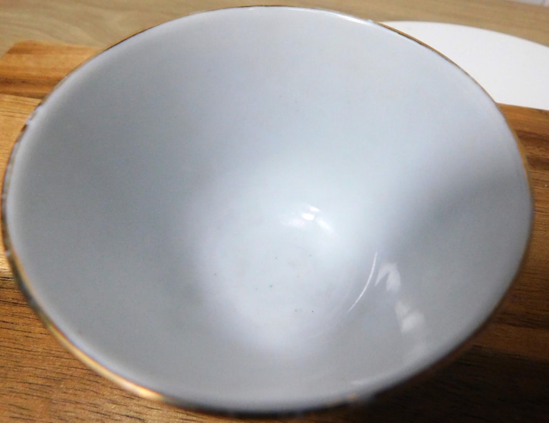 May include: A small, light gray bowl with a gold rim. The bowl is empty and appears to be made of ceramic. The bowl is sitting on a wooden surface.