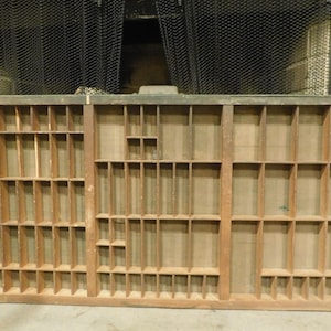 May include: A vintage wooden printer's tray with numerous small compartments of varying sizes. The tray is rectangular, with a light brown color and visible wood grain. It is likely used for organizing small items or displaying collectibles.