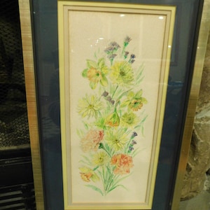 May include: A framed watercolor painting of a floral arrangement. The artwork features yellow, green, and orange flowers with purple accents. The painting is set against a light background, framed with a gold metal frame and a navy blue mat.