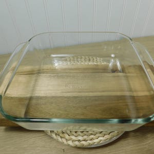 May include: A clear, square Pyrex baking dish with handles. The dish is made of glass and sits on a wooden cutting board. The background is a white wall with vertical wood paneling. The dish is approximately 9 inches square.