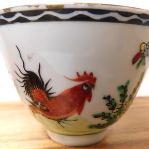 May include: White porcelain bowl with a hand-painted rooster design. The rooster is depicted in shades of red and orange, with black tail feathers. The bowl has a black and gold rim and a small butterfly and floral accents.