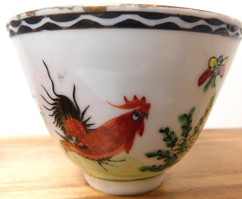 May include: White porcelain bowl with a hand-painted rooster design. The rooster is depicted in shades of red and orange, with black tail feathers. The bowl has a black and gold rim and a small butterfly and floral accents.