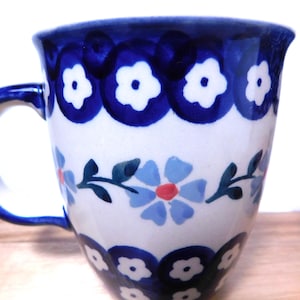 May include: A ceramic mug with a white base, adorned with blue floral patterns and a blue rim and handle. The design includes blue flowers with red centers and white flower-shaped accents. The mug is likely for beverages.