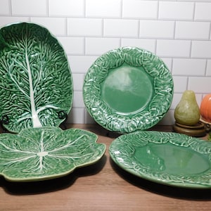 May include: A collection of four green ceramic plates and platters featuring embossed cabbage leaf textures. Two pieces are shaped like full cabbage heads with white veining, while two round plates feature detailed leaf borders and smooth centers.