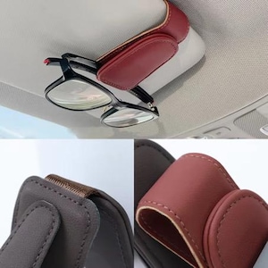 May include: A burgundy leather car visor glasses holder with a pair of black-framed eyeglasses clipped to it. The holder is attached to the car's interior roof. The holder is made of leather and has a stitched design.