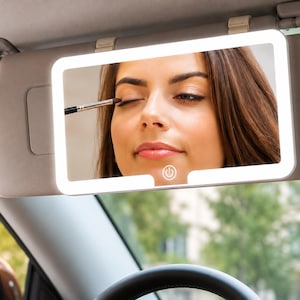 Puede incluir: Un espejo de cortesía rectangular para el parasol del coche con un borde de luz LED blanca y un botón de encendido táctil en el centro inferior. El espejo está montado en un parasol beige y refleja a una persona aplicándose maquillaje.
