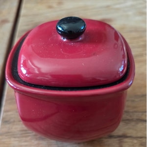 May include: A small, square-shaped red ceramic container with rounded corners and a matching red lid. The lid features a smooth, glossy finish and is topped with a small, round, black knob handle. The interior rim of the container is black.