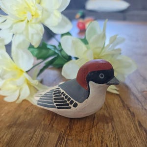 May include: A small ceramic bird figurine featuring a reddish-brown head, a black face mask, a grey back with black wing details, and a cream-coloured chest. The bird is positioned in front of several large, pale yellow artificial flowers.