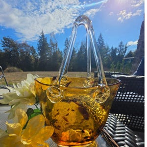 May include: A translucent amber-colored glass bowl shaped like a handbag, featuring two clear glass handles attached with decorative knotted glass accents. The bowl has a wide, open top and a smooth, rounded base, reflecting light through its warm, golden-toned glass surface.