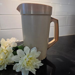 May include: A beige plastic jug with a matching lid and a sturdy handle, featuring a simple cylindrical body and a pouring spout. The jug is positioned next to two white artificial flowers with layered petals and green stems.