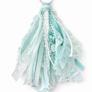 May include: A handmade tassel keychain featuring a mix of light blue and white ribbons, lace, and fabric strips. The tassel is adorned with decorative buttons and a silver metal ring for attachment. The overall design is soft and feminine.