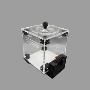 May include: A clear acrylic cube with a circular top and a black sphere handle. The cube has a white base and black hardware. A black and brown mechanism is attached to the side.