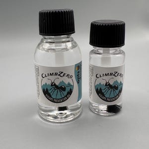 Puede incluir: Dos botellas de vidrio transparente con tapas negras. Cada botella tiene una etiqueta con el texto "CLIMBZERO ESCAPE PREVENTION OIL" y una ilustración de una hormiga. Las botellas están llenas de un líquido transparente.