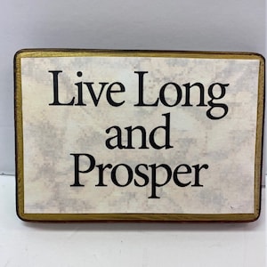 May include: A wooden sign with a gold border and a white background. The sign says "Live Long and Prosper" in black lettering.