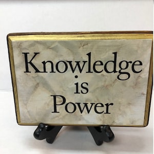 May include: A rectangular sign with the words "Knowledge is Power" in black lettering. The sign has a marble-like background and a gold-colored border. It is displayed on a black easel.