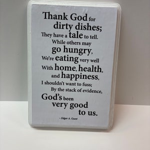 May include: A rectangular white sign with rounded corners features black text. The text reads, "Thank God for dirty dishes; They have a tale to tell...God's been very good to us." The author's name, Edgar A. Guest, is at the bottom.