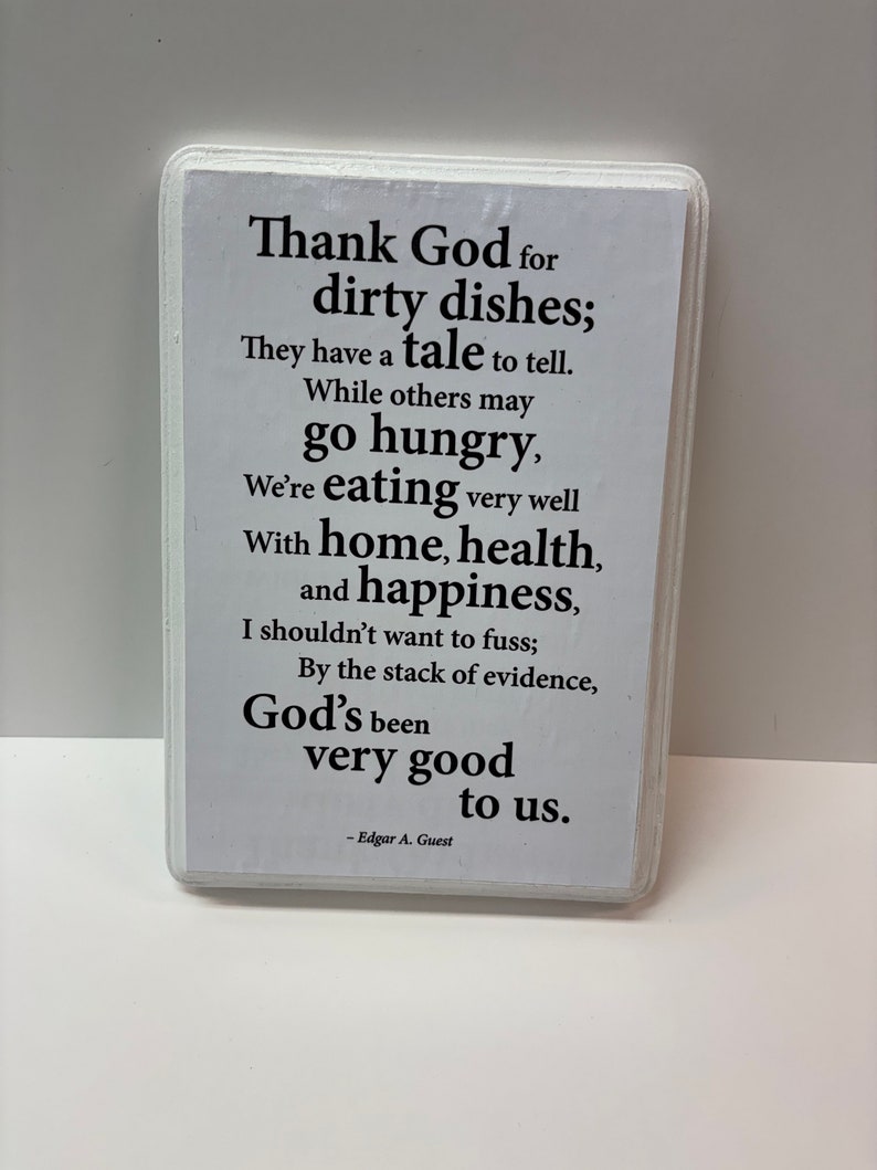 May include: A rectangular white sign with rounded corners features black text. The text reads, "Thank God for dirty dishes; They have a tale to tell...God's been very good to us." The author's name, Edgar A. Guest, is at the bottom.