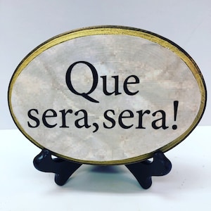 May include: A white oval wooden sign with a gold rim and a black stand. The sign says "Que sera, sera!"