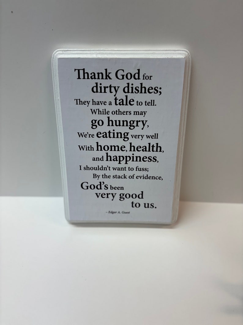 May include: A white rectangular plaque with rounded edges, featuring black text. The text reads, "Thank God for dirty dishes; They have a tale to tell...God's been very good to us." The plaque is a decorative item.