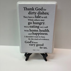 May include: A white rectangular sign with black text, displayed on a black easel. The sign features a poem about gratitude for dirty dishes, home, health, and happiness, attributed to Edgar A. Guest.